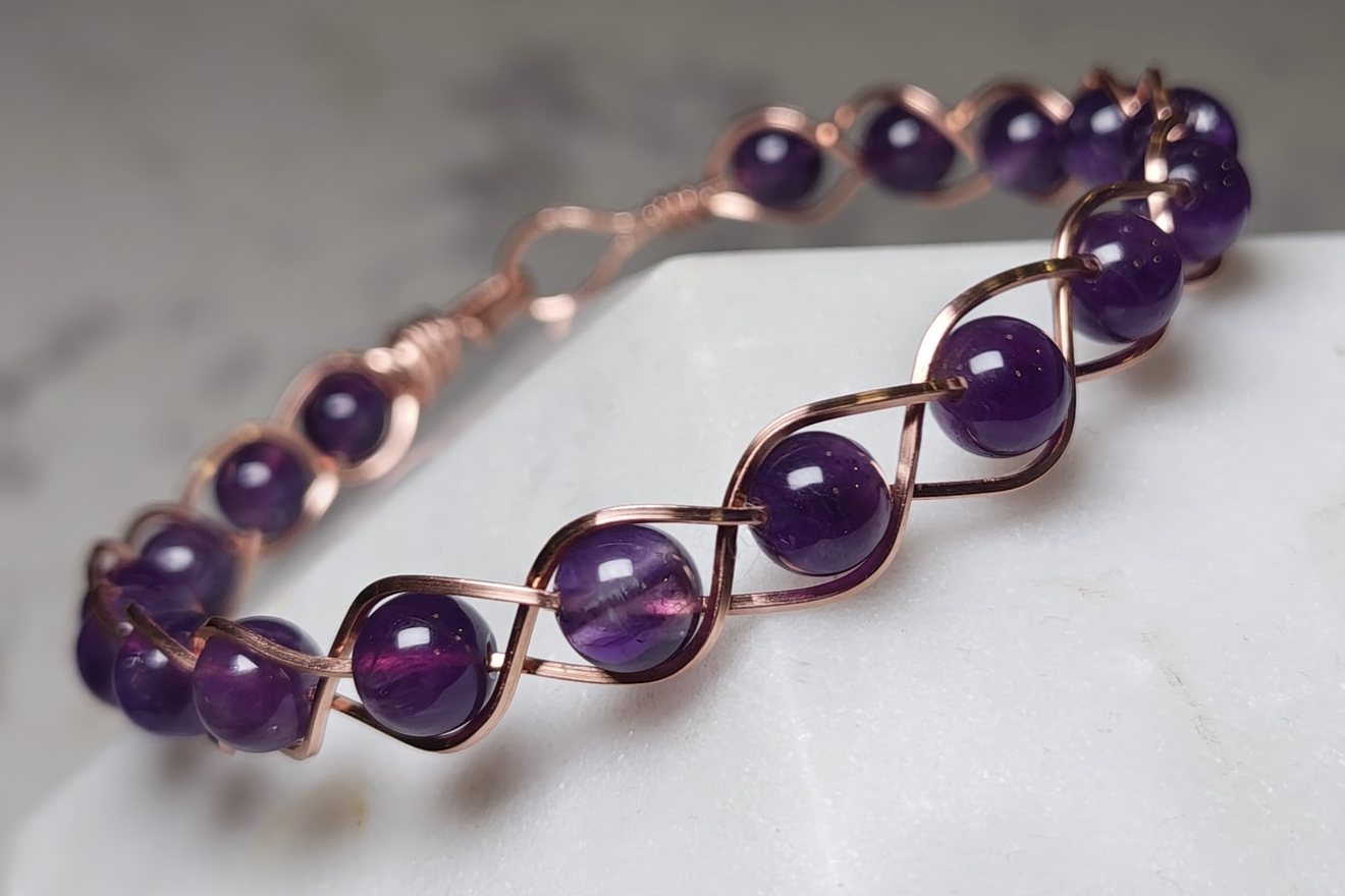 Bracelet of round amethyst beads held in a braided copper wire frame, resting on white stone.