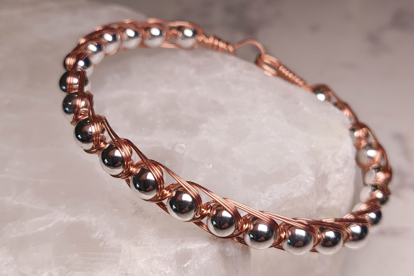 Bangle of polished hematite beads wrapped in fine copper wire, resting on a quartz block.